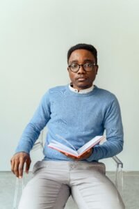 A thoughtful man in glasses and blue sweater holding a book indoors.